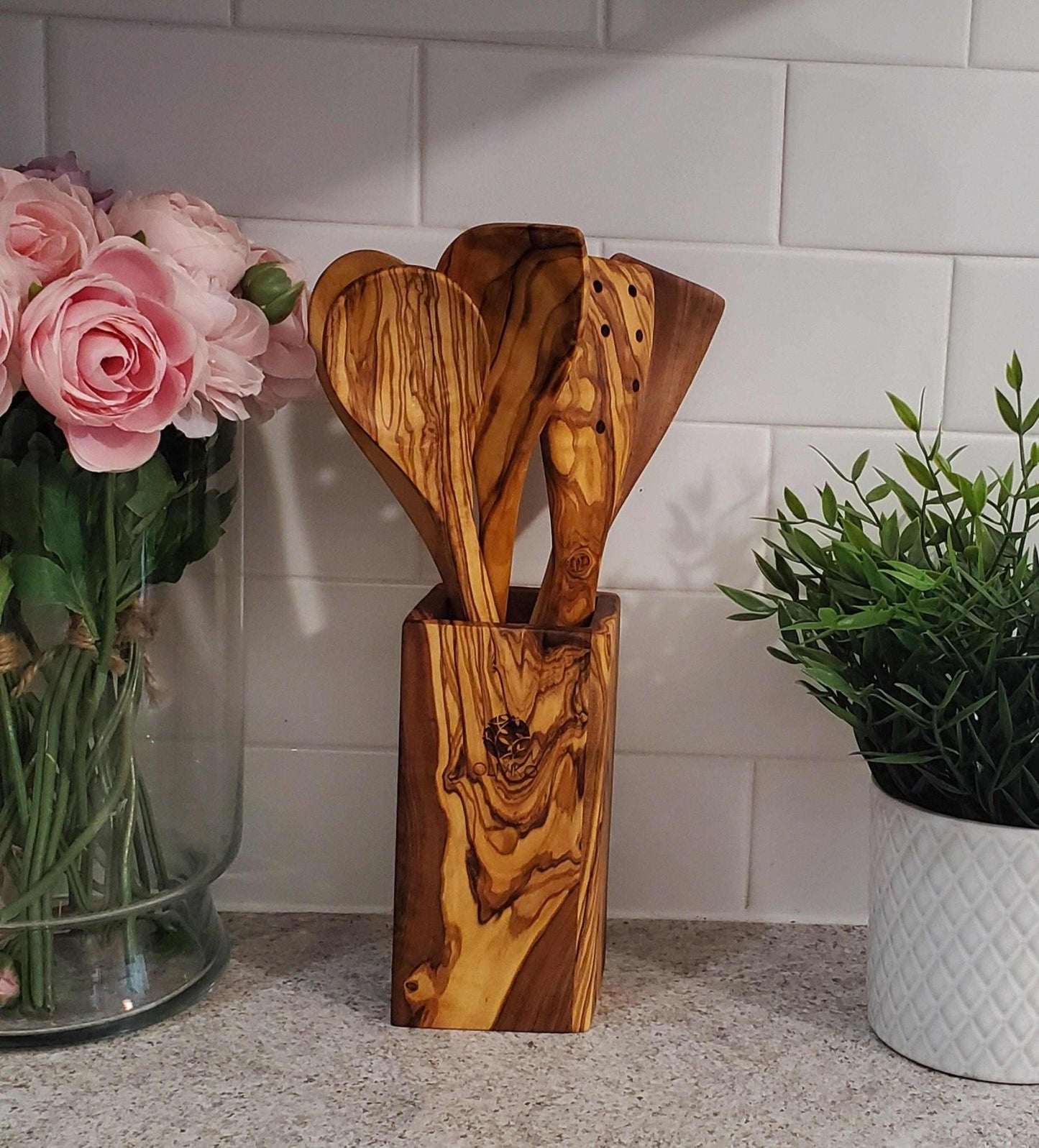 Handcrafted olive wood utensil set with holder, 2 spatulas and 3 spoons displayed on kitchen counter with flowers and plant nearby