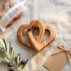 Two interlocking heart-shaped wooden baby teething rings with a natural olive wood grain pattern, displayed on a textured linen surface alongside an olive branch, a kraft paper tag, and a clear plastic pouch containing two additional rings.