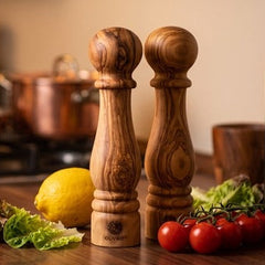 Two wooden salt and pepper mills standing on a dark wood kitchen countertop, with a lemon, lettuce, cherry tomatoes, and olive oil bottles in a warm, blurred kitchen background.