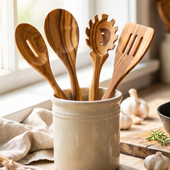 A lifestyle kitchen scene showing a person's hand using a wooden spoon to stir a black cast iron skillet filled with colorful vegetables on a wooden cutting board. A cream ceramic container holding various wooden utensils stands on the counter next to a window.