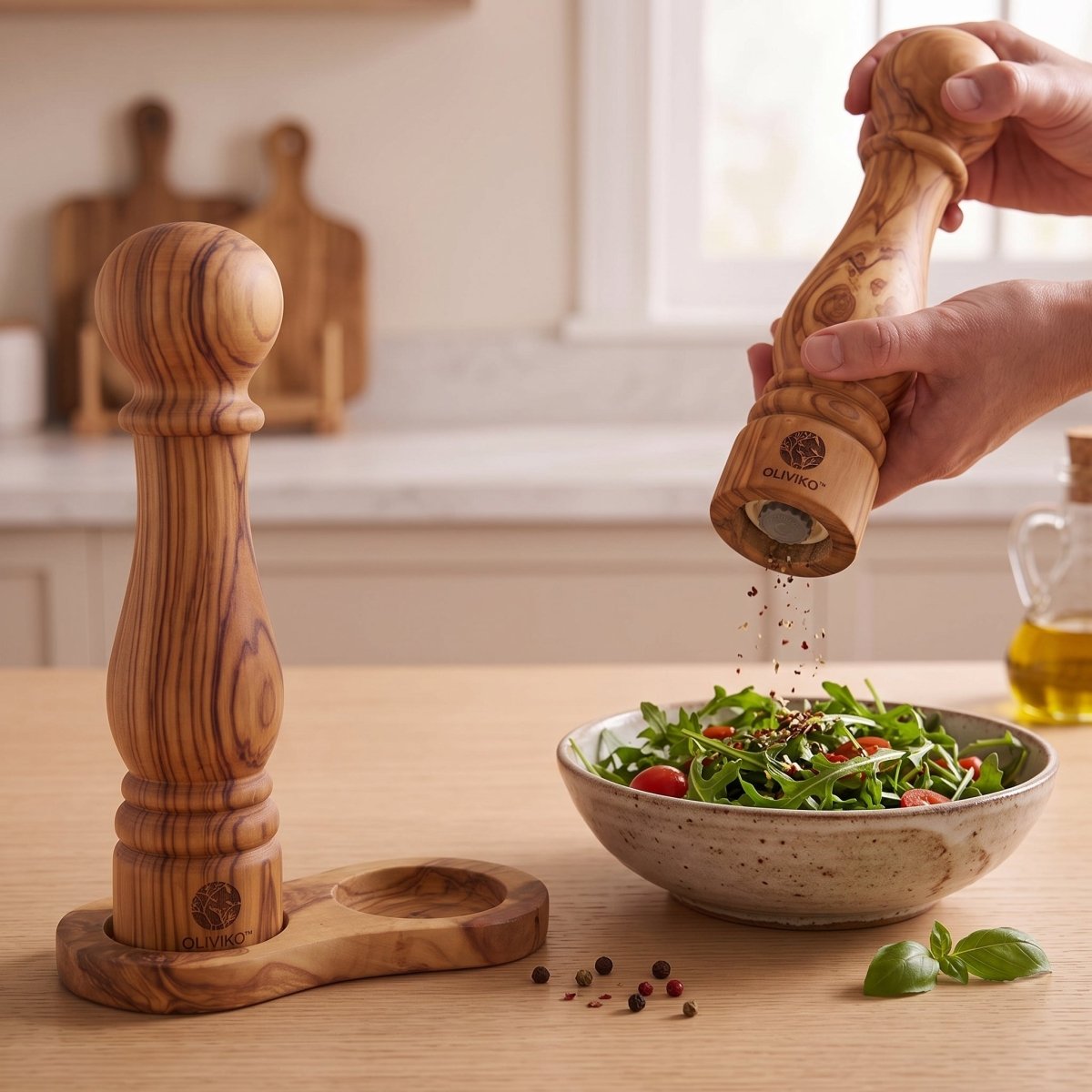 Two wooden pepper mills, one standing in a wooden holder and one being used by a hand to grind pepper over a salad bowl, set on a wooden table in a bright kitchen.