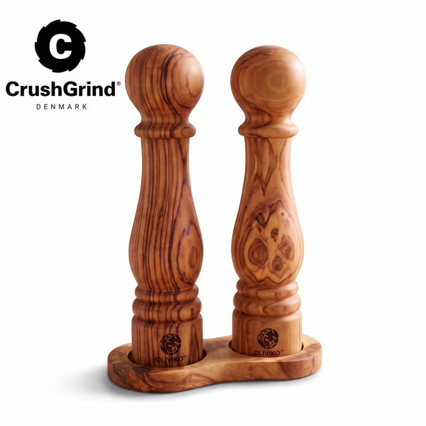 Two wooden pepper and salt mills with a rich grain pattern, standing in a matching wooden base on a white background. The CrushGrind logo is visible in the top left corner.